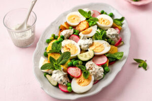 Angerichteter knuspriger Kartoffel-Ei-Salat auf einer weißen Platte, mit goldbraunen Kartoffeln, halbierten Eiern, Erbsen, Radieschen, Gurke, Minze und cremigem Joghurt-Kräuter-Dressing, daneben ein Glas Dressing mit Löffel