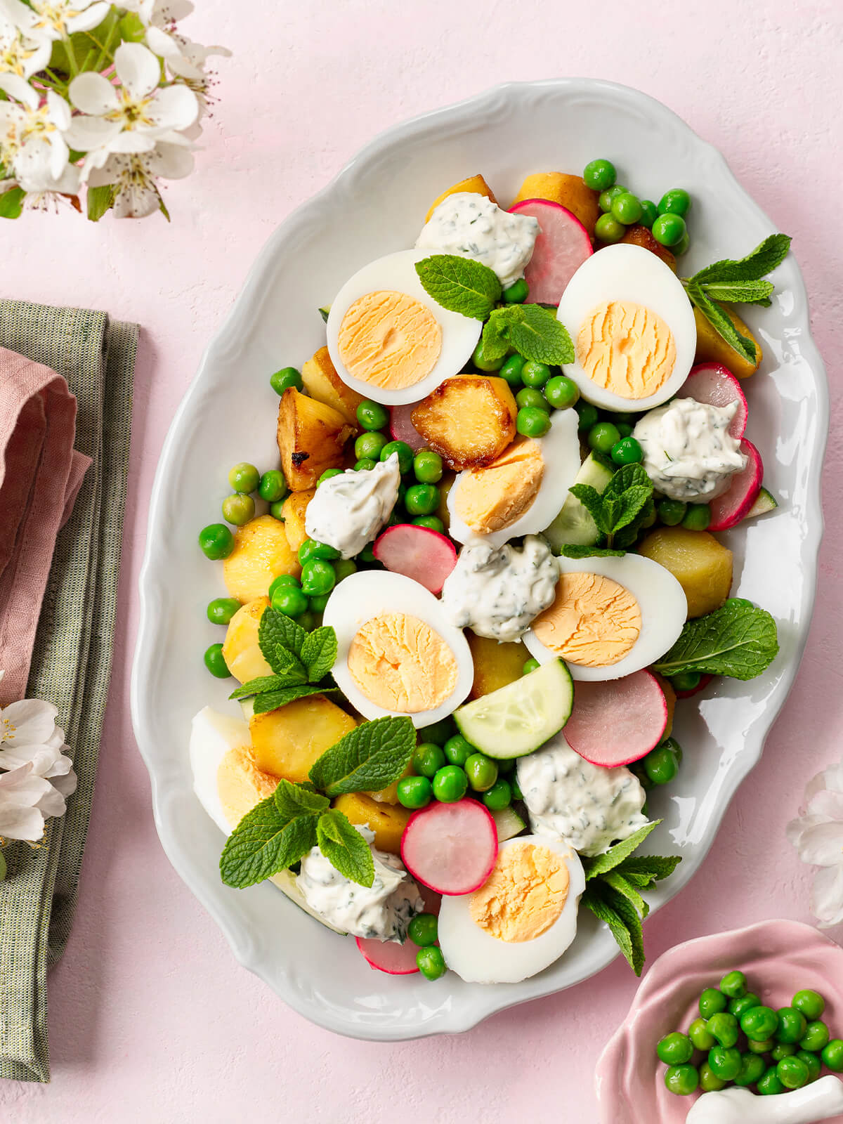 Knuspriger Kartoffel-Ei-Salat auf einer weißen Platte hochkant von oben, mit goldbraun gebratenen Kartoffeln, halbierten Eiern, grünen Erbsen, Radieschen, Gurke, viel frischer Minze und Klecksen cremigem Joghurt-Kräuter-Dressing, daneben Frühlingsblüten