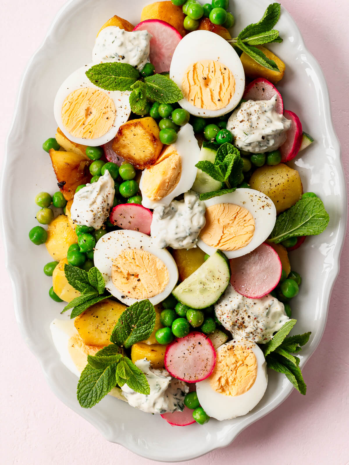 Nahaufnahme des knusprigen Kartoffel-Ei-Salats von oben, mit goldgelben, an den Kanten gebratenen Kartoffeln, halbierten Eiern mit festem gelbem Dotter, grünen Erbsen, Radieschen und Klecksen cremigem Joghurt-Kräuter-Dressing mit sichtbaren grünen Kräutern