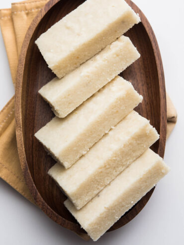 Weiße Kokos-Barfi-Riegel diagonal auf einem Holzteller angerichtet, von oben