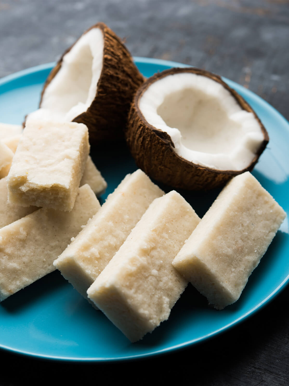 Nahaufnahme weißer Kokos-Barfi-Stücke auf einem türkisfarbenen Teller, daneben zwei Hälften einer frischen Kokosnuss