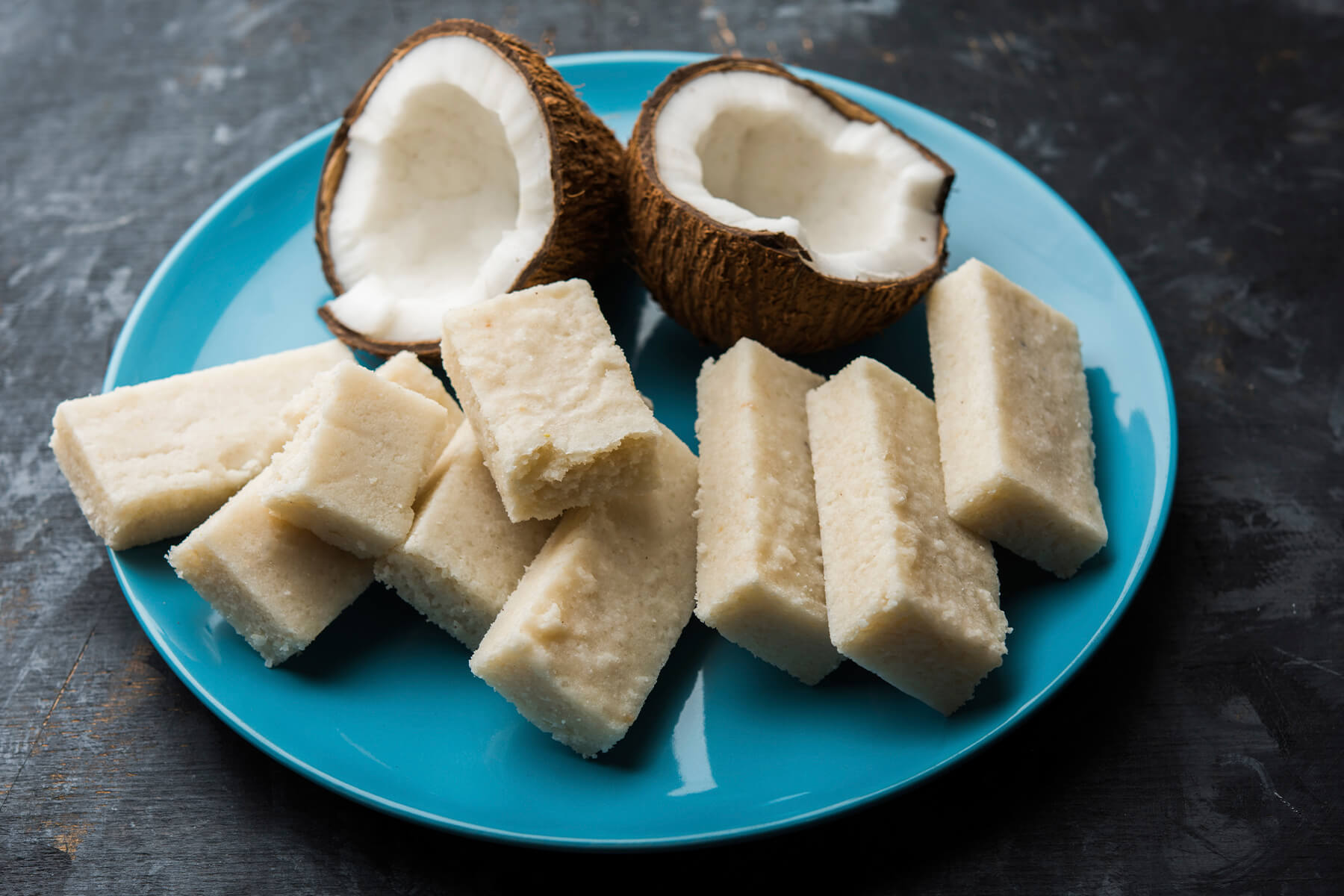 Weiße Kokos-Barfi-Stücke auf einem türkisfarbenen Teller mit zwei aufgeschlagenen Kokosnusshälften, auf dunklem Untergrund