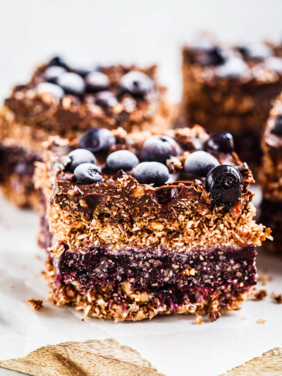 Vegane Kokos-Schoko-Schnitte im Anschnitt mit vier Lagen: Kokos-Hafer-Boden, Blaubeer-Chia-Schicht, Kokos-Hafer-Streusel und Schokoladenhaube mit gefrorenen Blaubeeren