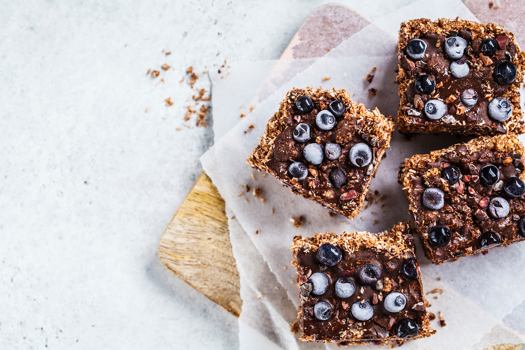 Vier vegane Kokos-Schoko-Schnitten mit Blaubeeren und Kakaonibs auf Backpapier von oben