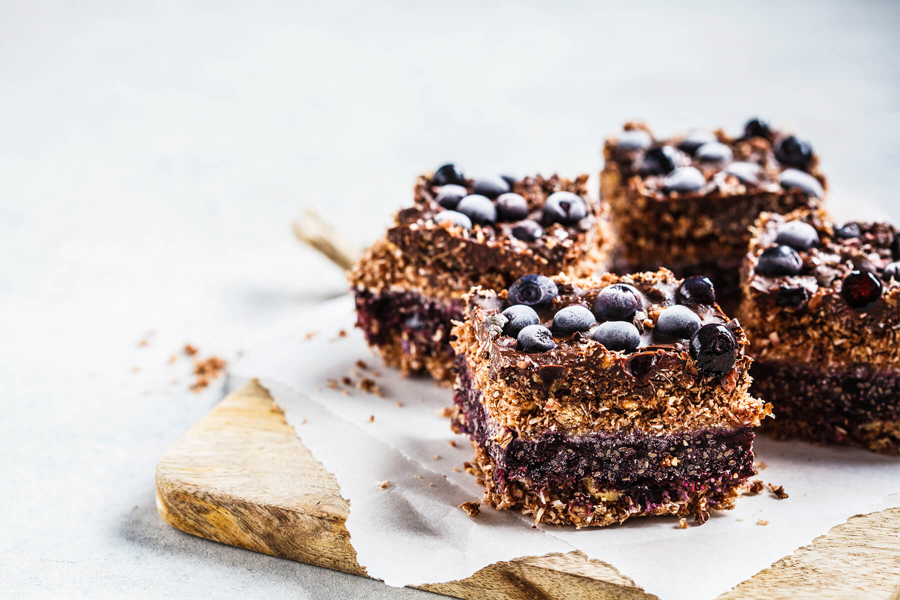 Nahaufnahme veganer Kokos-Schoko-Schnitten von der Seite mit vier erkennbaren Lagen und gefrorenen Blaubeeren