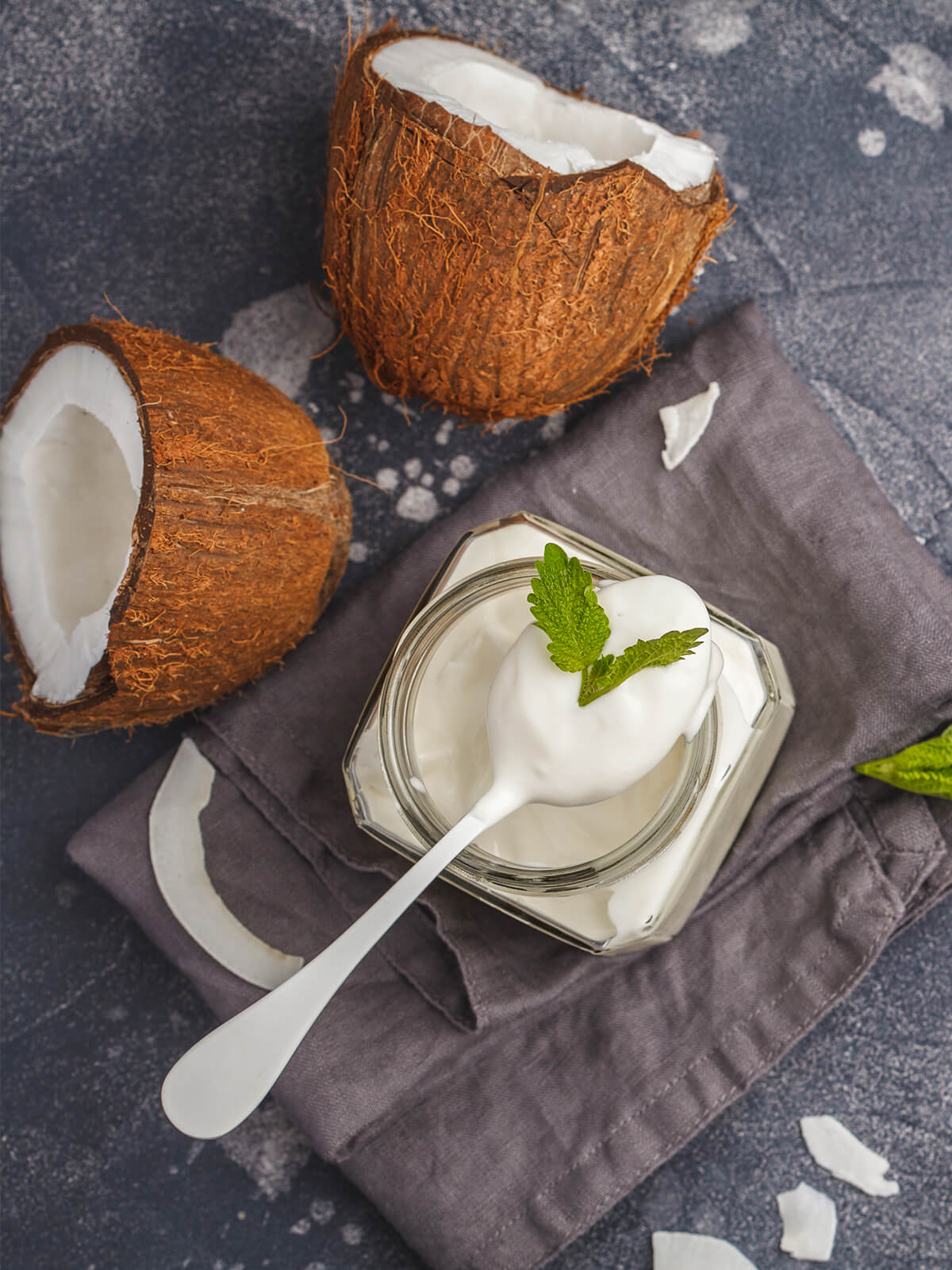 Cremiger veganer Kokosjoghurt im Glas mit Löffel und Minze, daneben aufgeschlagene Kokosnuss von oben