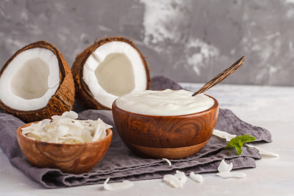 Cremiger veganer Kokosjoghurt in einer Holzschale mit Löffel, daneben Kokoshälften und Kokoschips