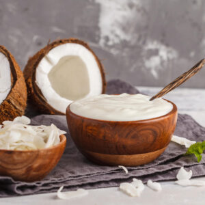 Cremiger veganer Kokosjoghurt in einer Holzschale mit Löffel, daneben Kokoshälften und Kokoschips