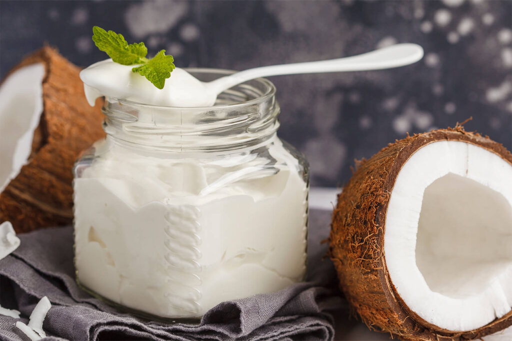 Nahaufnahme von cremigem Kokosjoghurt im Glas mit einem Löffel und frischer Minze, daneben eine Kokoshälfte