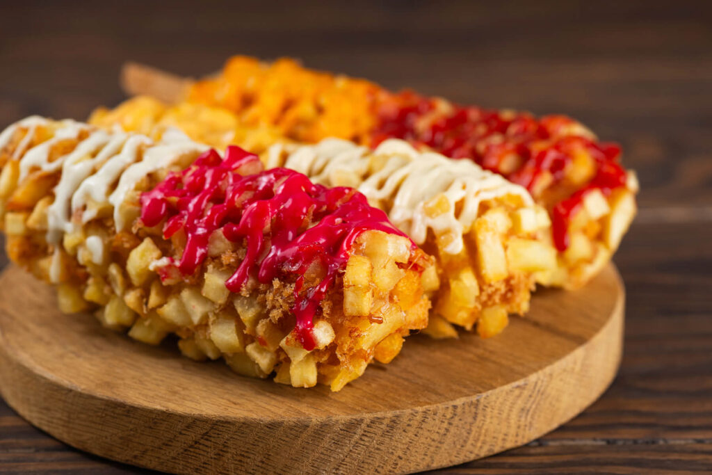 Drei koreanische Kartoffel-Corndogs auf einem runden Holzbrett, rundum mit hell-goldgelben Kartoffelwürfeln und Panko paniert und mit weißer, roter, käsegelber und pinkfarbener Beerensauce beträufelt