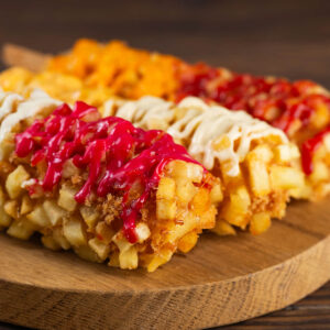 Drei koreanische Kartoffel-Corndogs auf einem runden Holzbrett, rundum mit hell-goldgelben Kartoffelwürfeln und Panko paniert und mit weißer, roter, käsegelber und pinkfarbener Beerensauce beträufelt