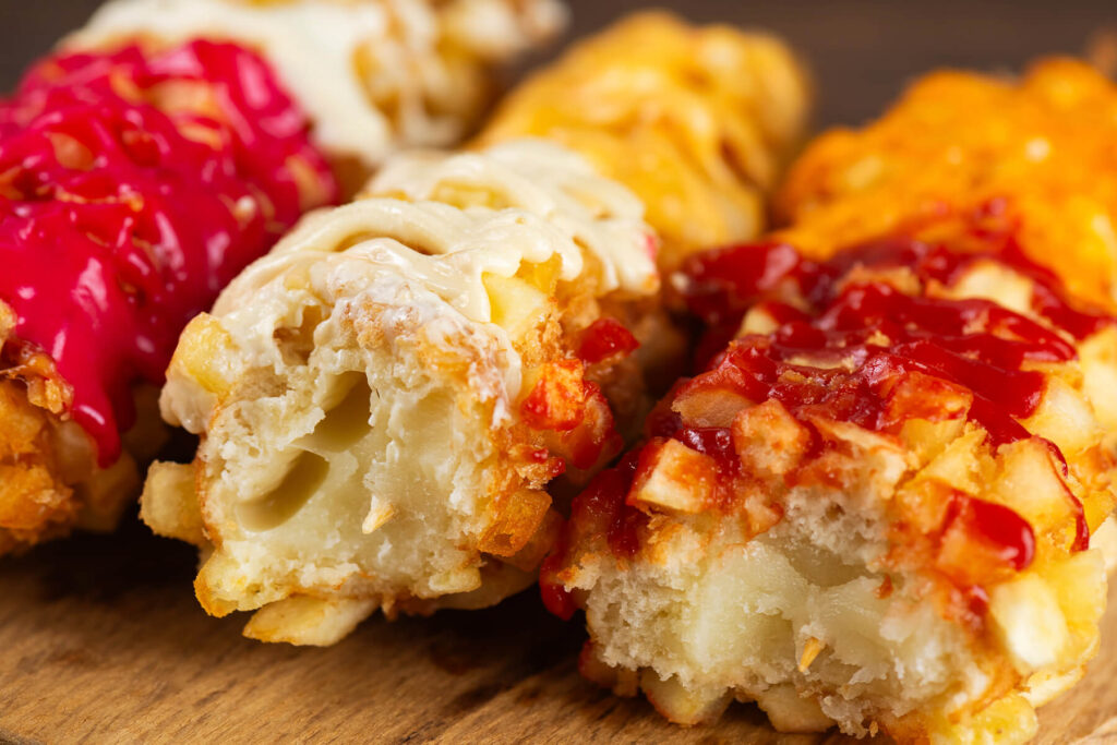 Angebissene koreanische Kartoffel-Corndogs auf einem Holzbrett im Anschnitt, im Kern der geschmolzene Mozzarella, darum eine dicke luftige Krume aus gewickeltem Hefeteig und außen die Kruste aus Kartoffelwürfeln und Panko mit weißer, roter und pinkfarbener Sauce