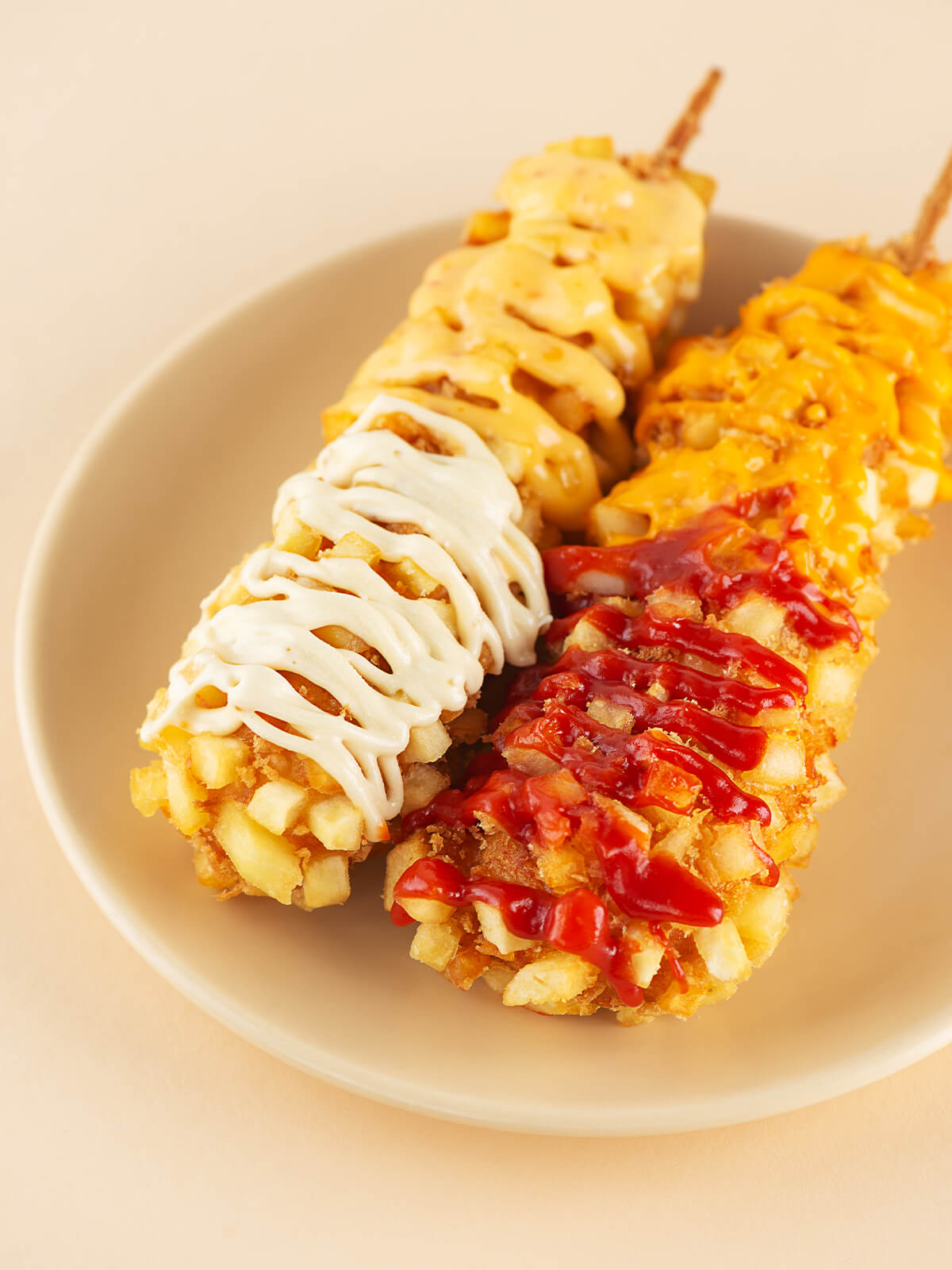 Zwei koreanische Kartoffel-Corndogs am Holzspieß auf einem beigen Teller, rundum mit hell-goldgelben Kartoffelwürfeln und Panko paniert und mit weißer, käsegelber und roter Sauce beträufelt