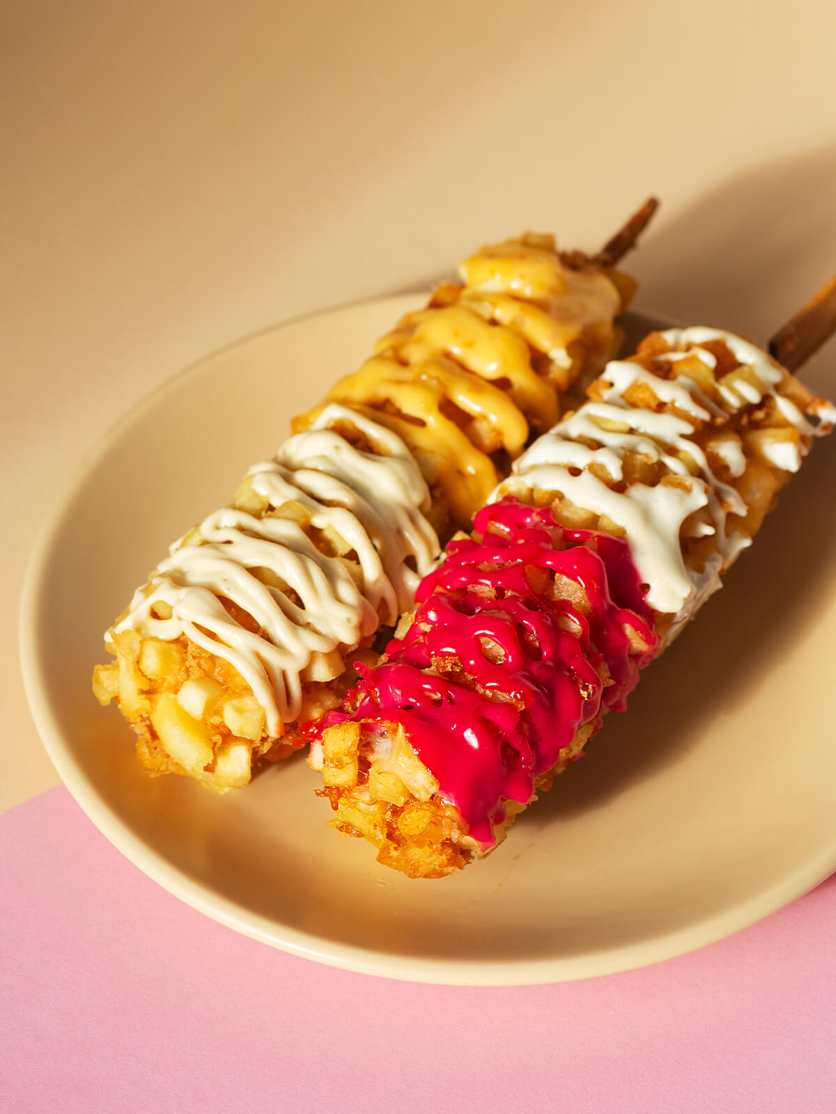 Zwei koreanische Kartoffel-Corndogs am Holzspieß auf einem beigen Teller vor rosa Hintergrund, paniert mit Kartoffelwürfeln und Panko und beträufelt mit weißer, käsegelber und pinkfarbener Sauce