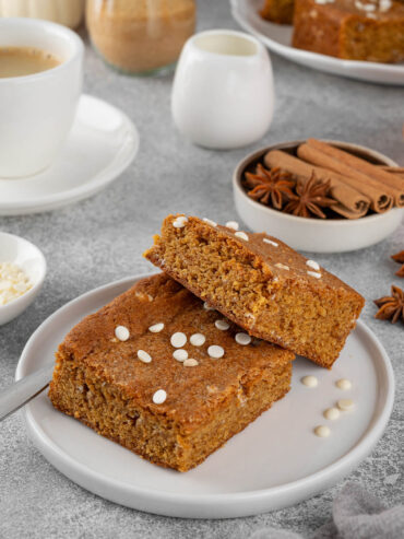 Zwei Kürbis-Blondies auf einem weißen Teller mit weißen Schokoladentropfen, dahinter eine Tasse Kaffee und Zimtstangen