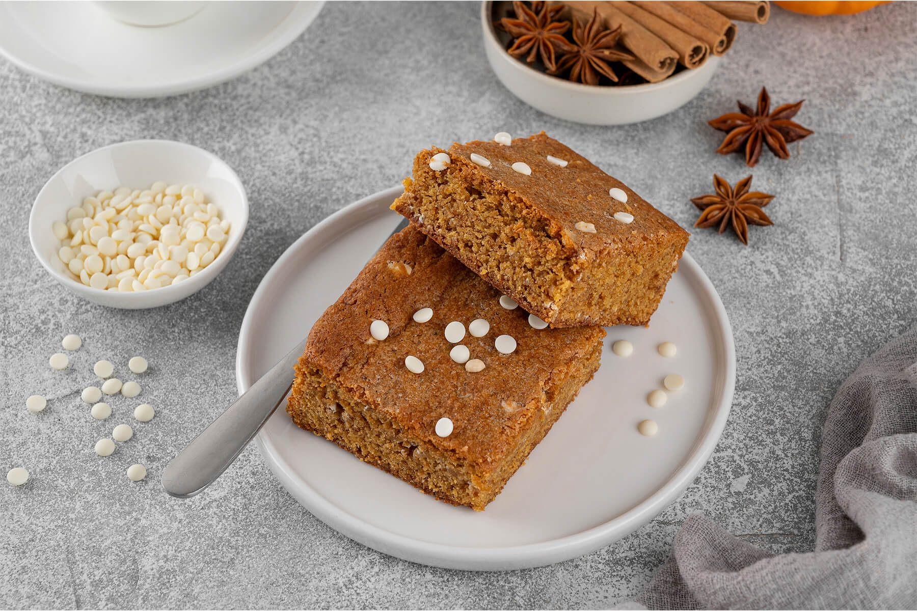 Zwei Kürbis-Blondies auf einem hellen Teller, daneben eine Schale mit weißen Schokoladentropfen, dazu Sternanis und Zimtstangen