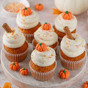 Kürbis-Cupcakes mit weißem Frischkäse-Frosting, Fondant-Mini-Kürbissen und Zimtstangen auf einem hellen Holzbrett
