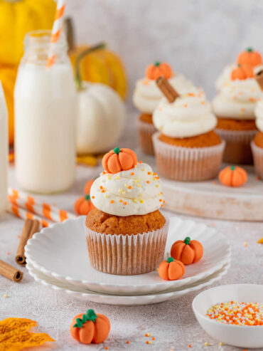 Kürbis-Cupcake mit hoher Frischkäse-Frosting-Haube und Fondant-Kürbis auf einem weißen Teller, dahinter Milchflasche