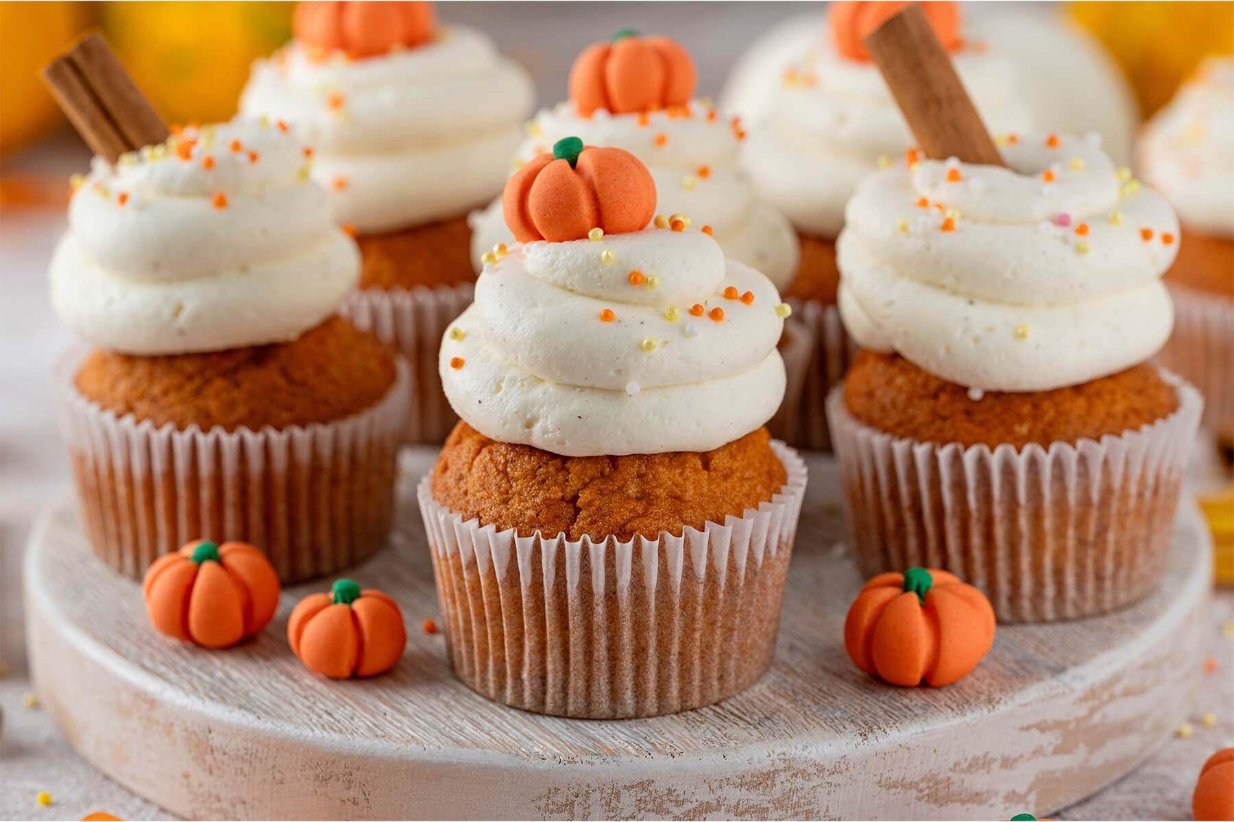 Mehrere Kürbis-Cupcakes mit Frischkäse-Frosting, Zimtstangen und Fondant-Kürbissen nebeneinander auf hellem Holz