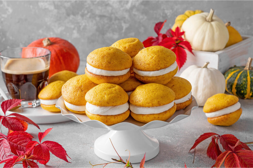 Weiche Kürbis-Whoopie-Pies mit Frischkäse-Creme auf einer weißen Etagere, dahinter Kaffee sowie weiße und orange Zierkürbisse