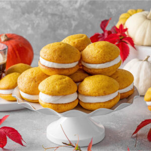 Weiche Kürbis-Whoopie-Pies mit Frischkäse-Creme auf einer weißen Etagere, dahinter Kaffee sowie weiße und orange Zierkürbisse