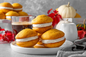 Mehrere fertige Kürbis-Whoopie-Pies mit hervorquellender Frischkäse-Creme, teils gestapelt, auf einem weißen Teller, dazu ein Glas Kaffee