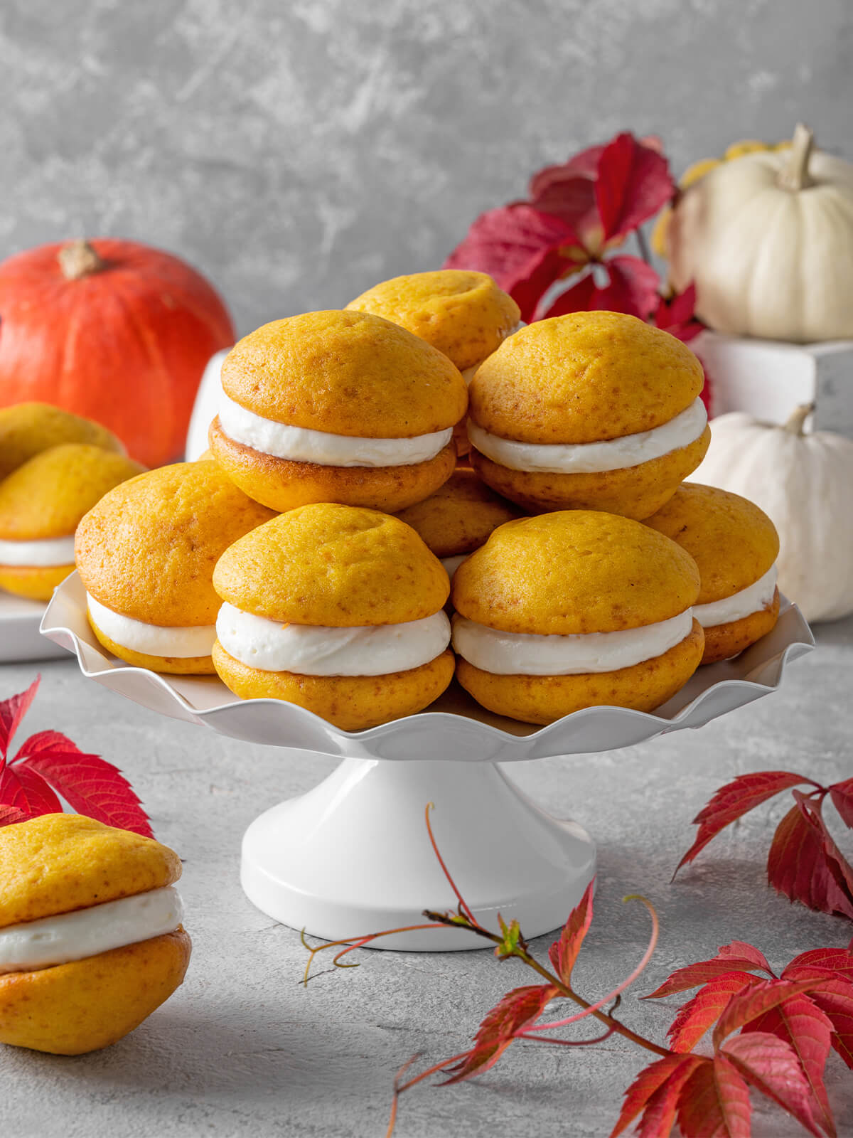 Gestapelte weiche Kürbis-Whoopie-Pies mit dicker Frischkäse-Creme auf einer weißen Etagere, dekoriert mit Herbstlaub und Kürbissen