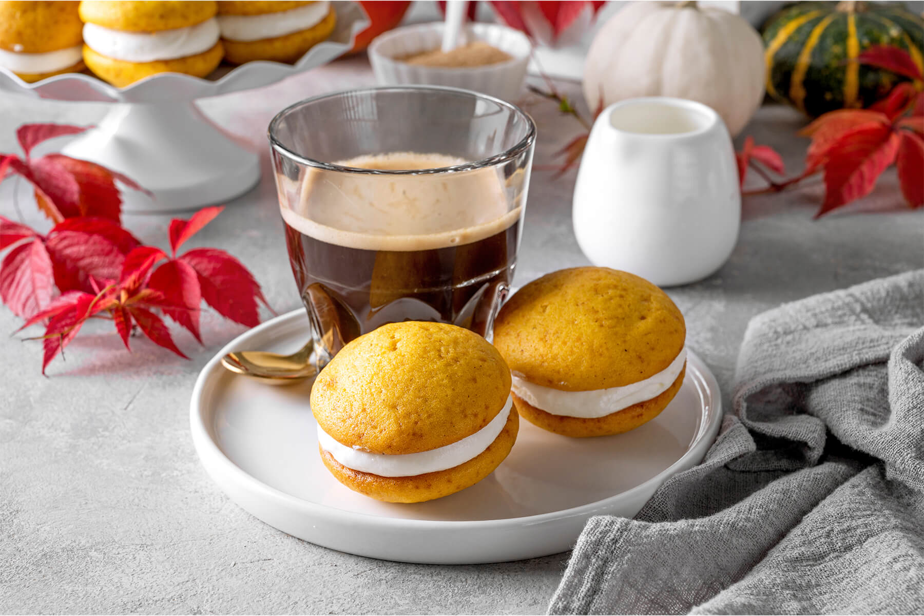 Zwei Kürbis-Whoopie-Pies mit dicker Frischkäse-Creme nebeneinander auf einem weißen Teller, dahinter ein Glas Kaffee