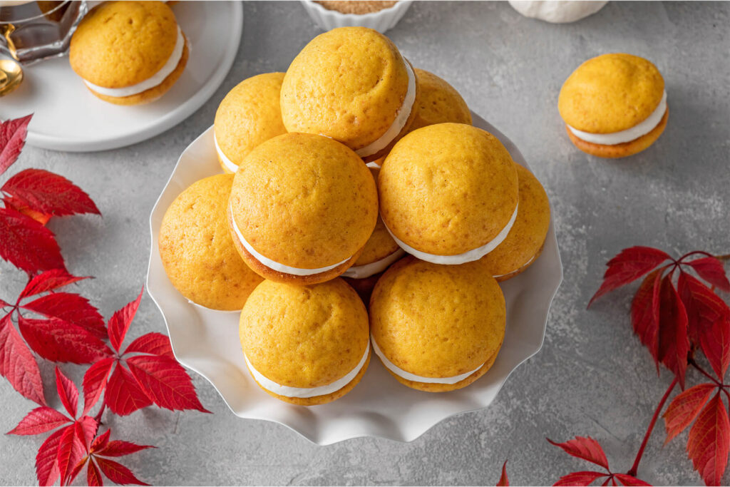 Kürbis-Whoopie-Pies von oben auf einer gewellten weißen Etagere, umgeben von rotem Herbstlaub