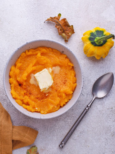 Cremiger Kürbisbrei mit schmelzender Butter und Zimt in einer gesprenkelten Schale, daneben ein dekorativer Zierkürbis und ein Herbstblatt