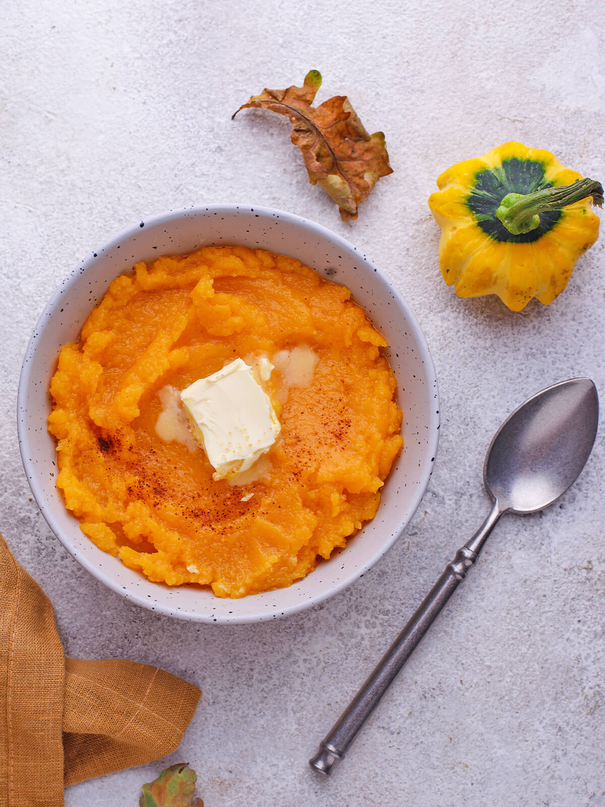 Cremiger Kürbisbrei mit schmelzender Butter und Zimt in einer gesprenkelten Schale, daneben ein dekorativer Zierkürbis und ein Herbstblatt