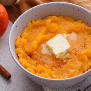 Cremiger Kürbisbrei mit schmelzender Butter und Zimt in einer gesprenkelten Schale, daneben ein Hokkaido-Kürbis und Zimtstangen