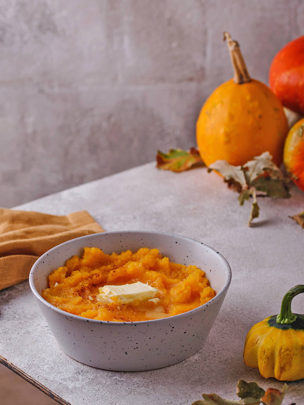 Schale mit Kürbisbrei, Butter und Zimt auf einem hellen Tisch, im Hintergrund zwei ganze Kürbisse und Herbstlaub, daneben eine ockerfarbene Serviette