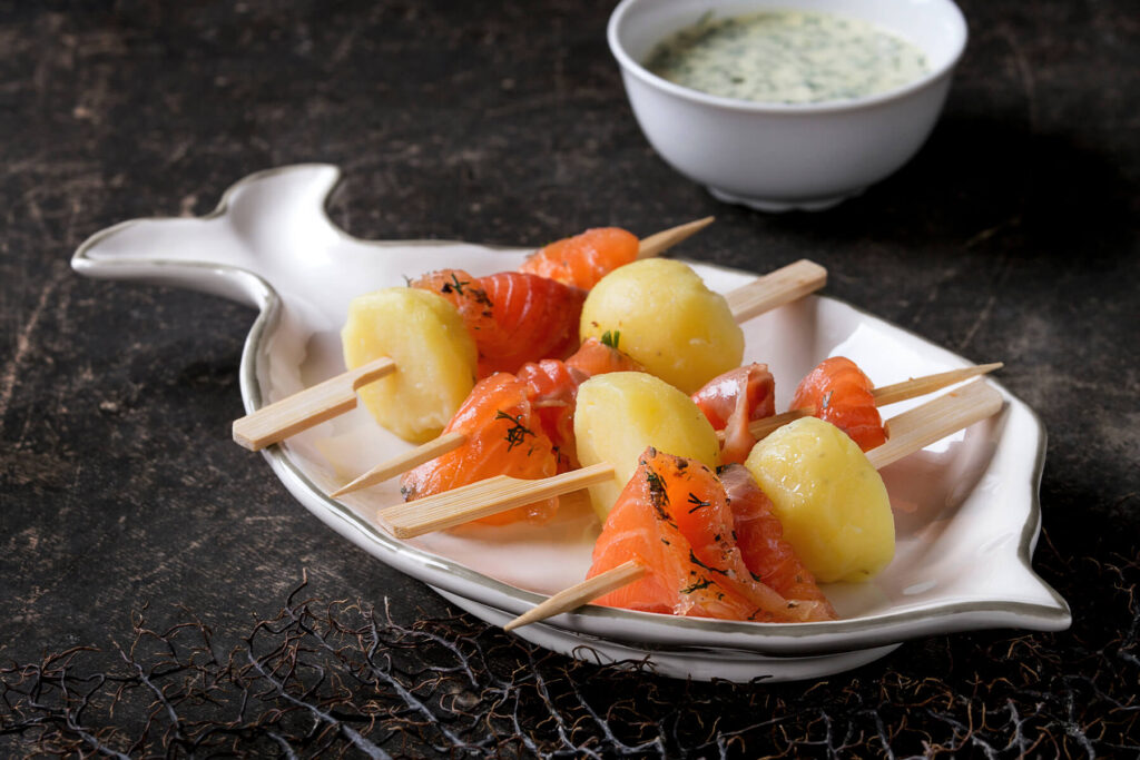 Mehrere flache Holzspieße mit je einer kleinen geschälten gekochten Kartoffel und einem gefalteten Stück kräftig orangerotem gebeiztem Lachs mit Dill und grobem Pfeffer, angerichtet auf einem weißen Servierteller in Fischform, im Hintergrund eine weiße Schale mit heller, grün gesprenkelter Kräutersauce auf dunklem Steinuntergrund