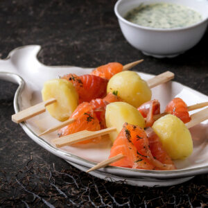 Mehrere flache Holzspieße mit je einer kleinen geschälten gekochten Kartoffel und einem gefalteten Stück kräftig orangerotem gebeiztem Lachs mit Dill und grobem Pfeffer, angerichtet auf einem weißen Servierteller in Fischform, im Hintergrund eine weiße Schale mit heller, grün gesprenkelter Kräutersauce auf dunklem Steinuntergrund