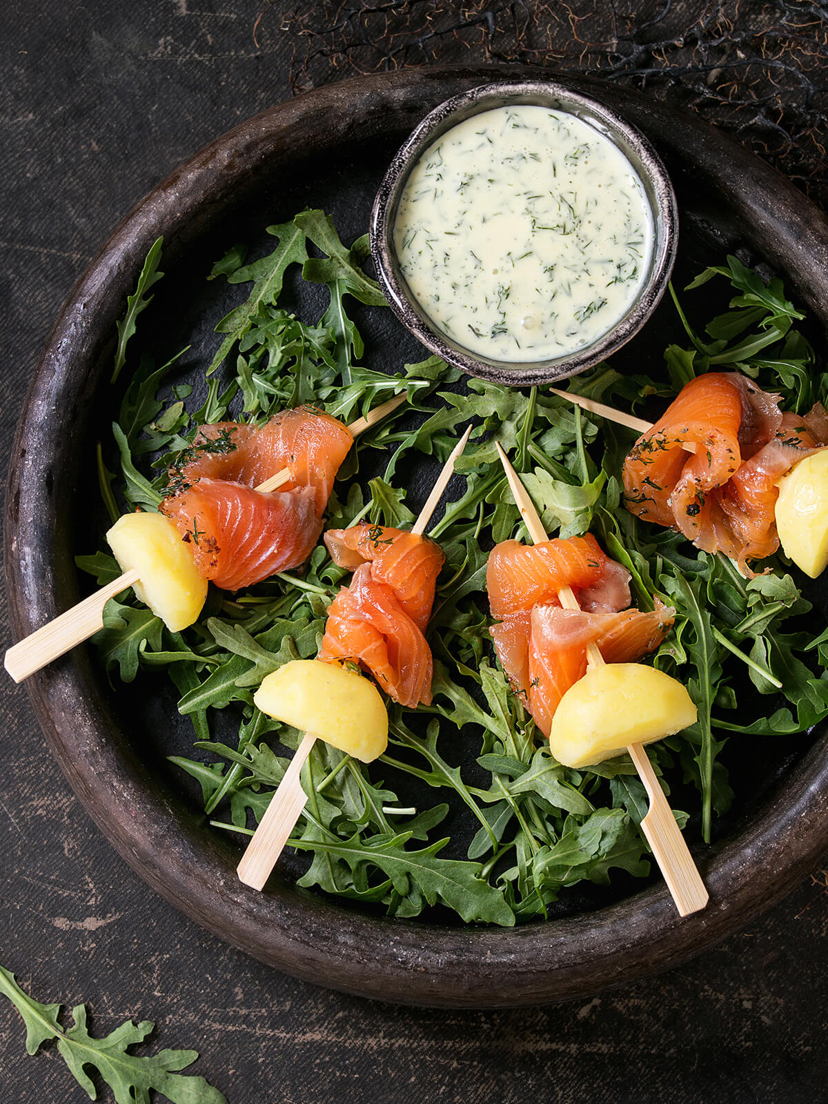 Vier Holzspieße mit je einem gefalteten Stück gebeiztem Lachs und einem Stück gekochter Kartoffel auf einem Bett aus frischem Rucola in einer dunklen runden Schale, dazu ein kleines Schälchen mit heller, cremig-fließender Kräutersauce mit fein verteiltem Dill, dunkler Untergrund