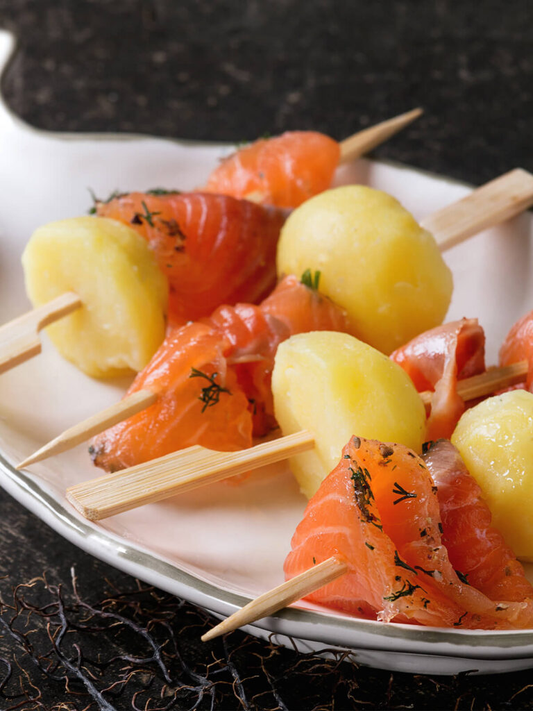 Nahaufnahme mehrerer Spieße auf einem weißen Servierteller in Fischform: kräftig orangerote Stücke gebeizter Lachs mit feinem Dill und grob gemörsertem Pfeffer, dazwischen kleine geschälte Kartoffeln mit glänzender Oberfläche, flache Holzspieße mit breitem Griffende, dunkler Hintergrund