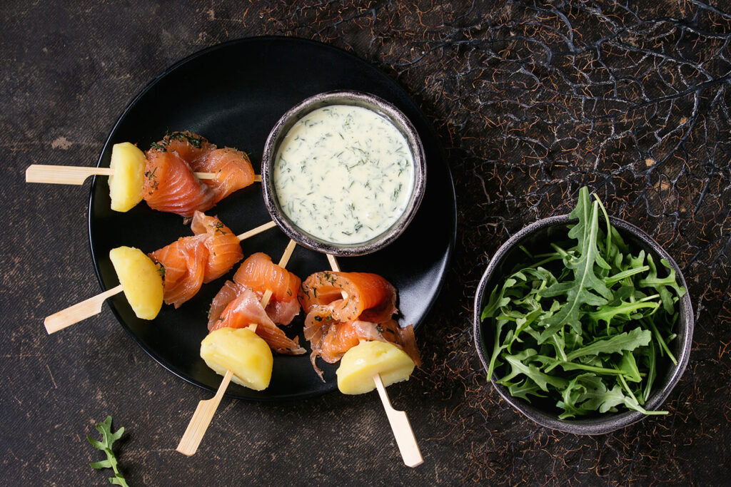Vier fertige Lachs-Kartoffel-Spieße von oben auf einem schwarzen runden Teller: gefaltete Stücke gebeizter Lachs mit Dill und ein halbiertes gelbes Kartoffelstück je Spieß, daneben ein kleines Schälchen mit heller, cremig-fließender Kräutersauce mit fein verteiltem Dill und eine dunkle Schale mit frischem Rucola auf dunklem Untergrund