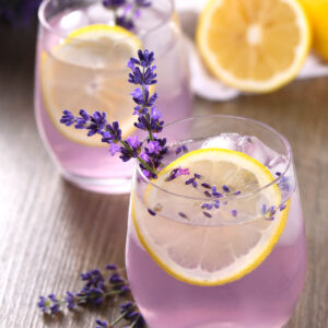 Zwei Gläser lila Lavendel-Blaubeer-Limonade mit Zitronenscheiben, Lavendelrispen und Eiswürfeln, daneben frische Zitronen