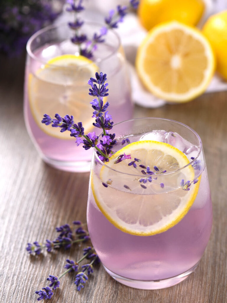Zwei Gläser lila Lavendel-Blaubeer-Limonade mit Zitronenscheiben, Lavendelrispen und Eiswürfeln, daneben frische Zitronen