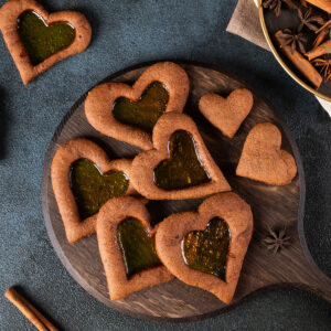 Lebkuchen-Herzen mit bernsteinfarbenem Bonbon-Fenster auf rundem Holzbrett, dazu Zimtstangen und Sternanis