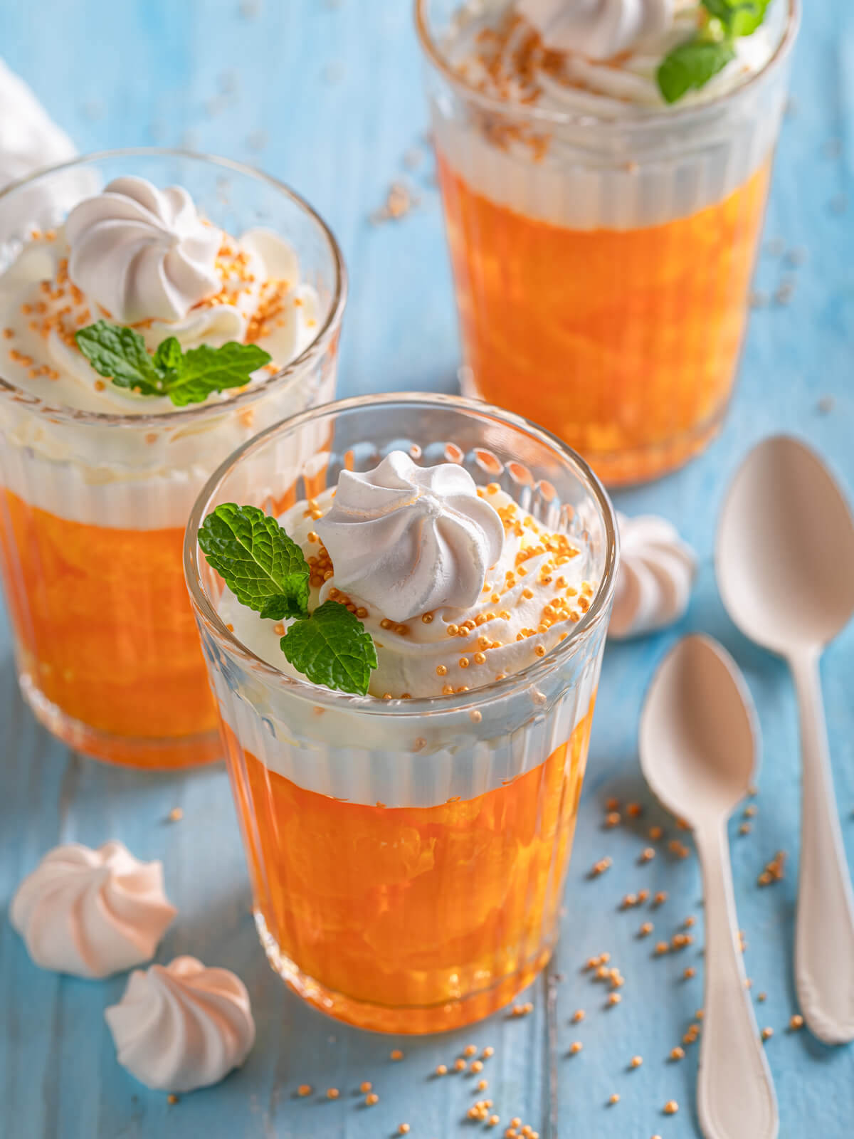 Klares Mandarinen-Gelee im Glas mit aufgespritzter Sahne, Baiser-Küsschen, goldenen Zuckerperlen und Minzblatt von schräg oben