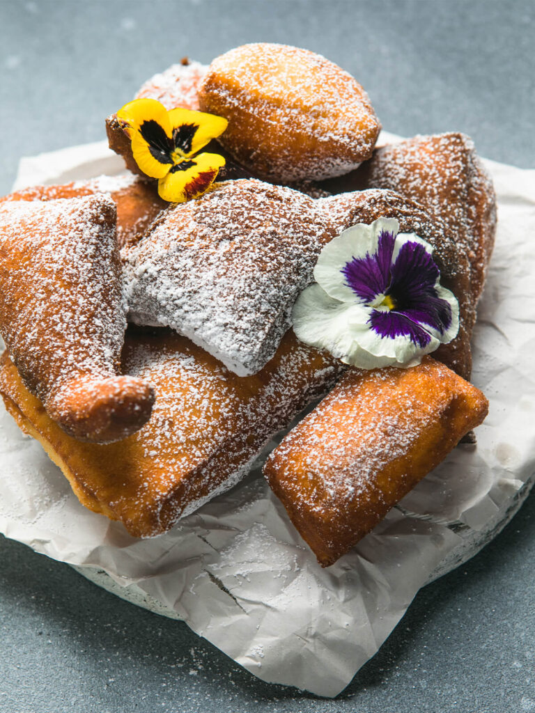 Goldbraune Mandazi mit Puderzucker bestäubt, angerichtet mit essbaren Blüten auf Backpapier