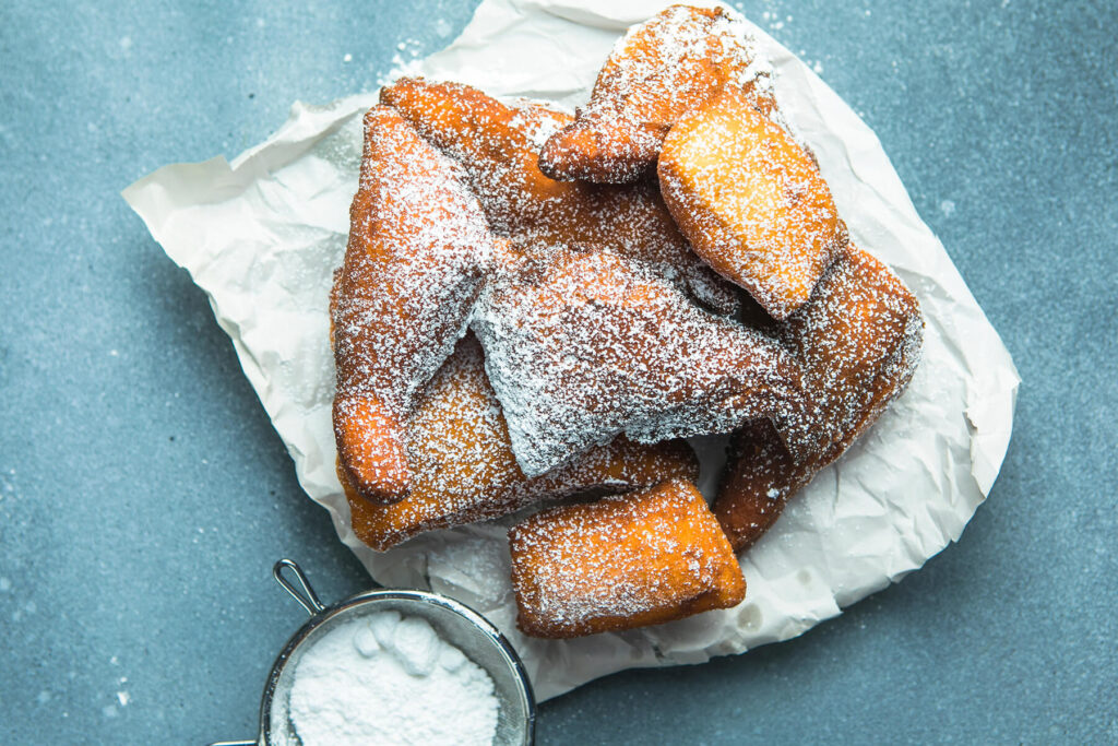 Goldbraune Mandazi mit Puderzucker bestäubt auf Backpapier, daneben ein Sieb mit Puderzucker