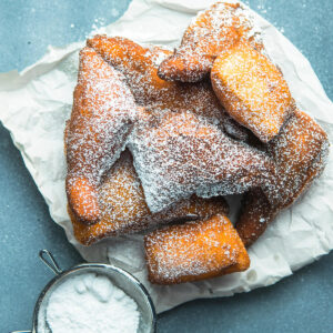 Goldbraune Mandazi mit Puderzucker bestäubt auf Backpapier, daneben ein Sieb mit Puderzucker