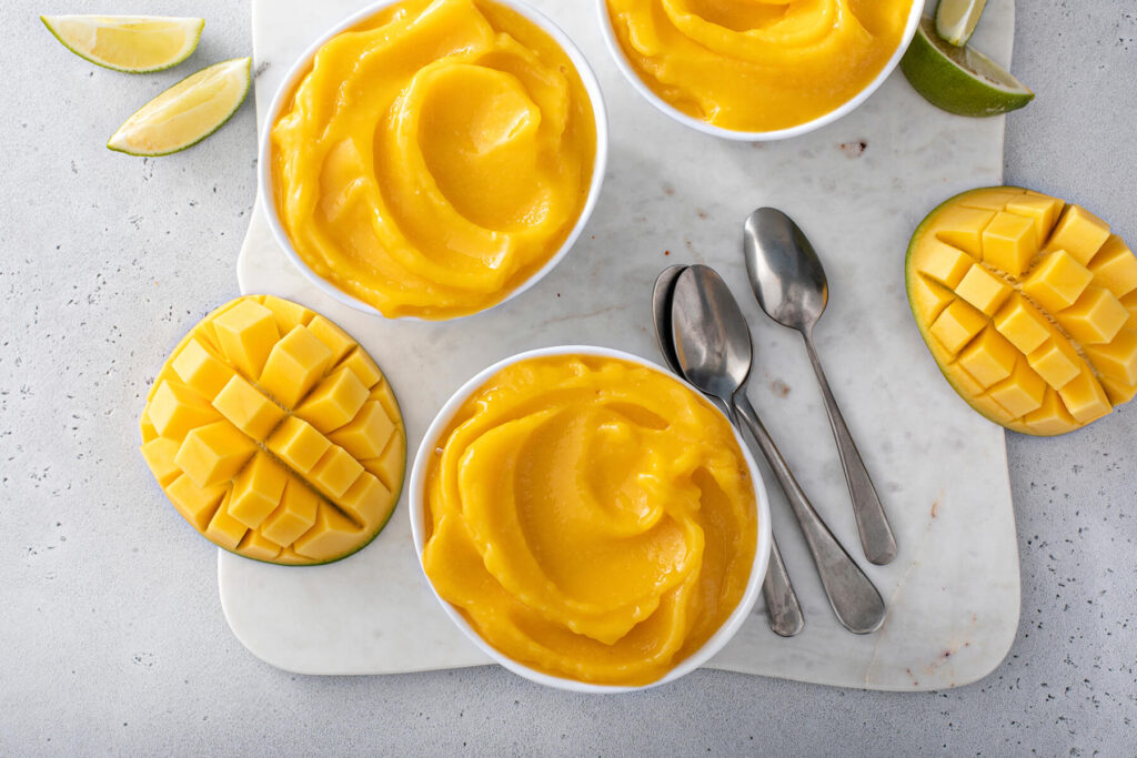 Mehrere weiße Schälchen mit cremiger, leuchtend gelb-oranger Mango Nice Cream von oben, dazu Limettenspalten, aufgeschnittene Mango im Igel-Schnitt und zwei Löffel auf einem hellen Marmorbrett