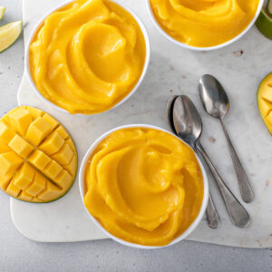 Mehrere weiße Schälchen mit cremiger, leuchtend gelb-oranger Mango Nice Cream von oben, dazu Limettenspalten, aufgeschnittene Mango im Igel-Schnitt und zwei Löffel auf einem hellen Marmorbrett