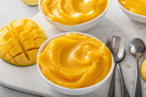 Nahaufnahme eines weißen Schälchens mit cremiger, weich geschwungener Mango Nice Cream in leuchtendem Gelb-Orange, daneben frische Mango im Igel-Schnitt und zwei Löffel
