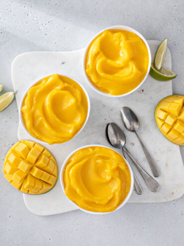 Mehrere weiße Schälchen mit cremiger, leuchtend gelb-oranger Mango Nice Cream von oben, dazu Limettenspalten und aufgeschnittene Mango im Igel-Schnitt auf einem hellen Marmorbrett