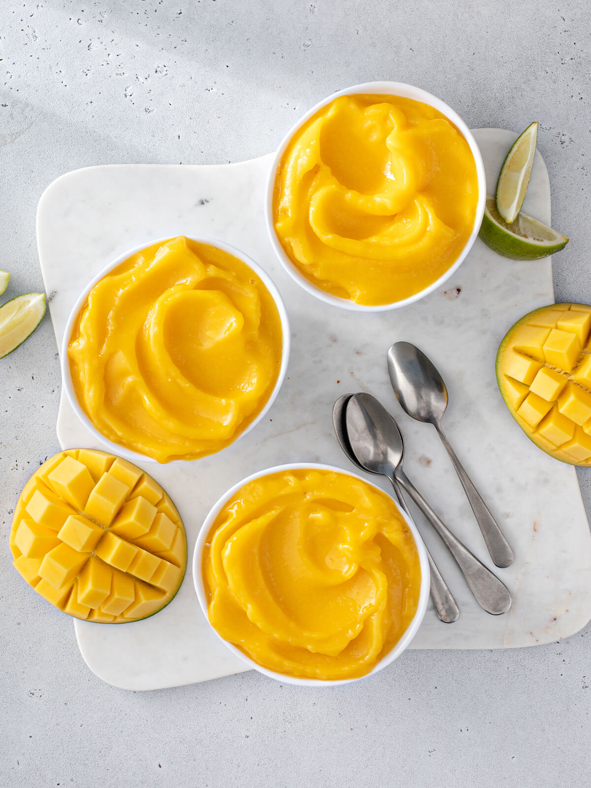 Mehrere weiße Schälchen mit cremiger, leuchtend gelb-oranger Mango Nice Cream von oben, dazu Limettenspalten und aufgeschnittene Mango im Igel-Schnitt auf einem hellen Marmorbrett